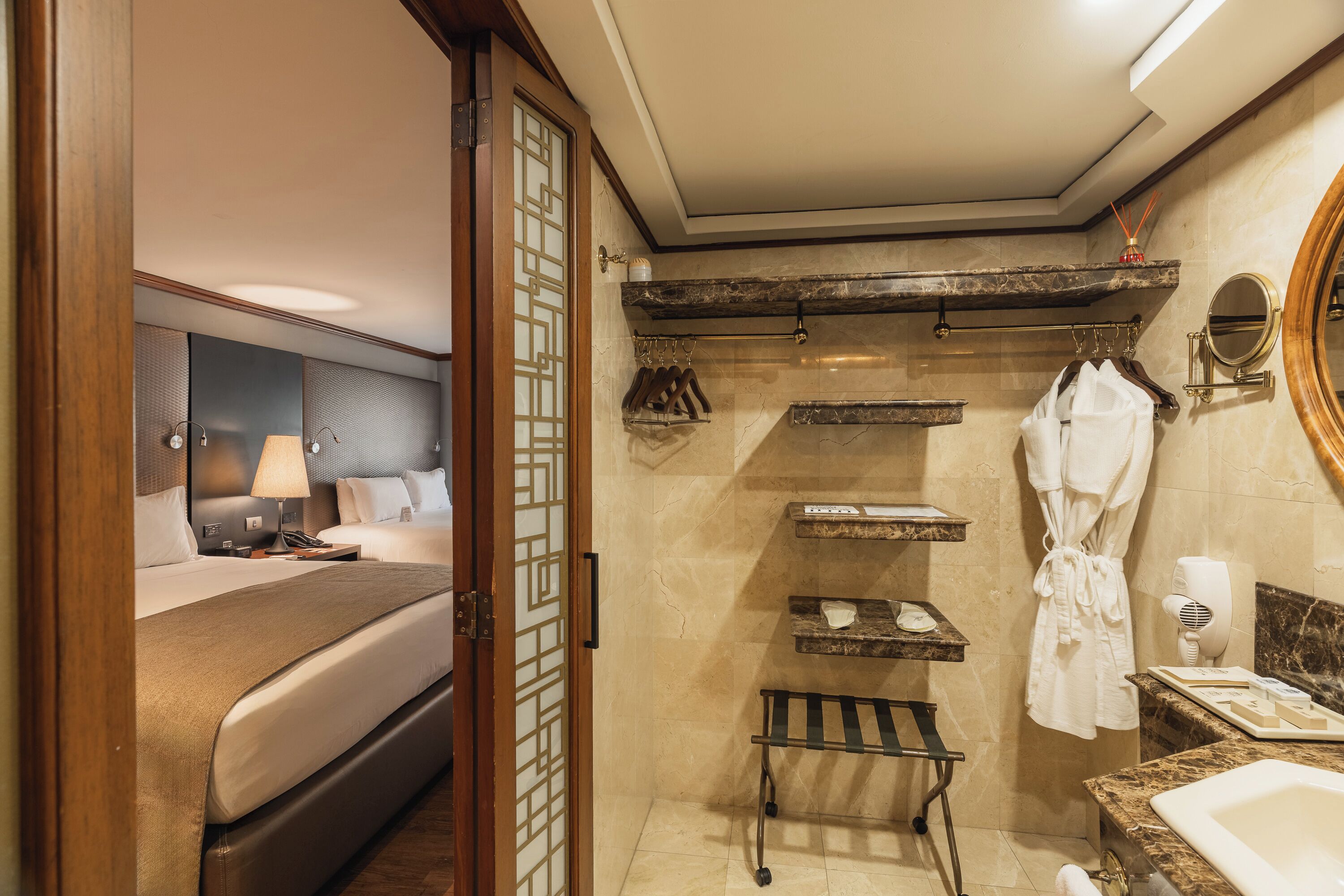 A hotel room with a bed, bedside table, lamp, and a bathroom with a sink, mirror, robe, and towel rack.