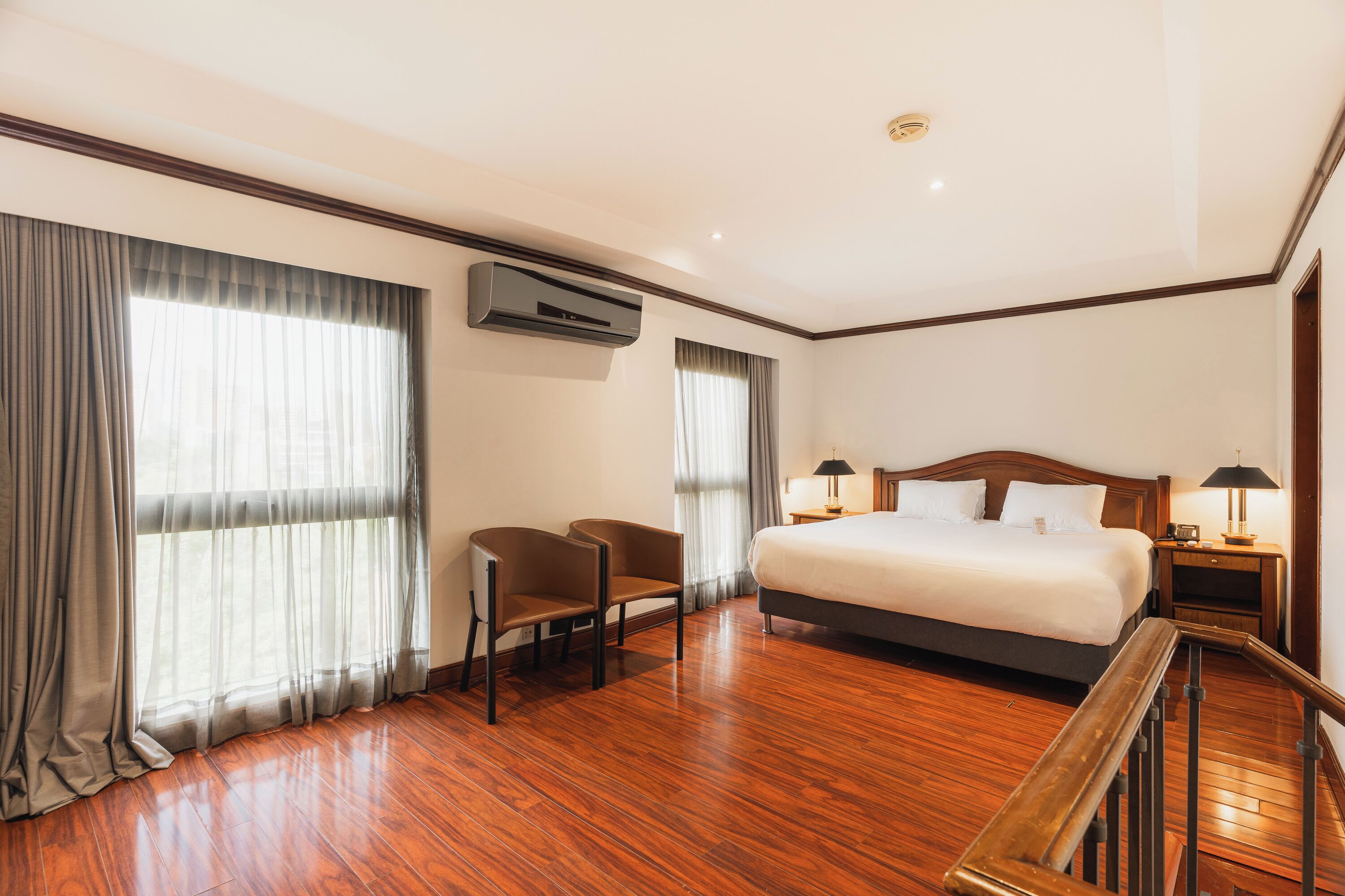 A hotel room with a large bed, two chairs, a desk, a nightstand, and an air conditioning unit.