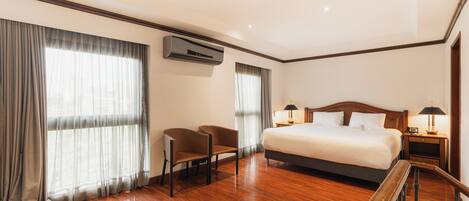 A hotel room with a large bed, two chairs, a desk, a nightstand, and an air conditioning unit.
