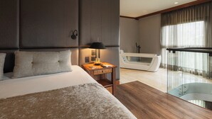 A modern hotel room with a large bed, a bedside table with a lamp, a glass-enclosed balcony, and a bathtub.