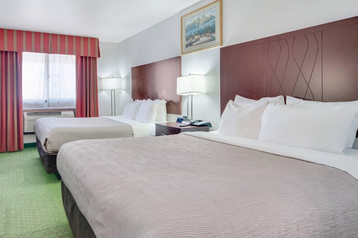 A hotel room with two beds, a large window with curtains, a painting on the wall, and a bedside lamp.