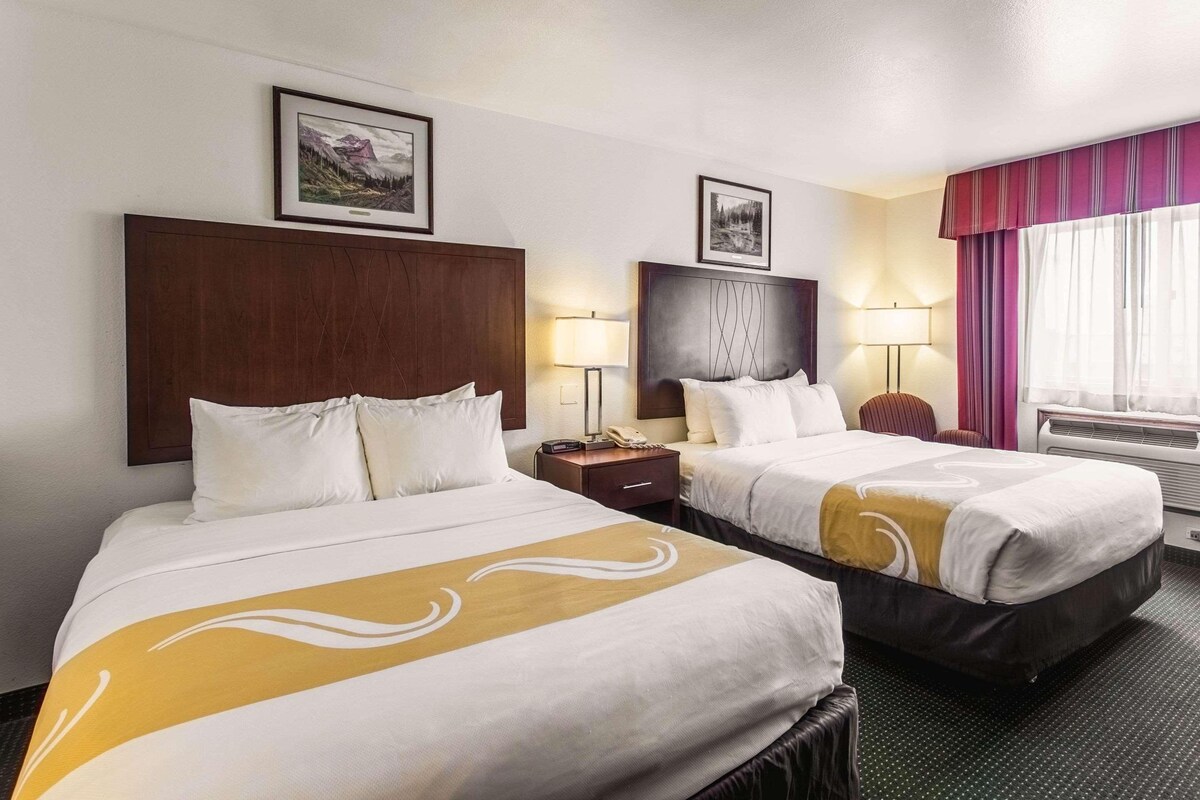 A hotel room with two beds, each with a wooden headboard, a nightstand, a lamp, and a window with curtains.