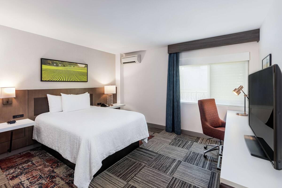A hotel room with a large bed, a desk with a flat-screen TV, a chair, a desk lamp, a wall-mounted air conditioner, and a window with curtains.