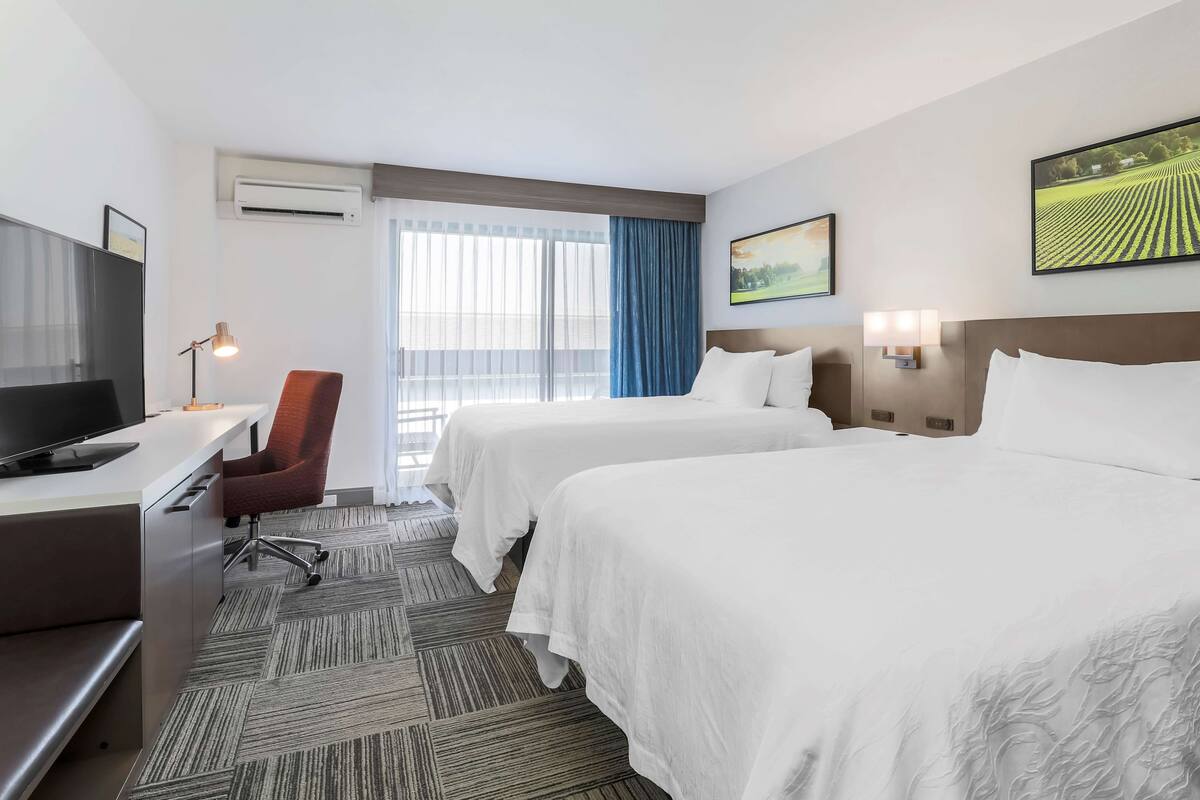 A hotel room with two beds, a desk with a TV, a chair, and a window with curtains.