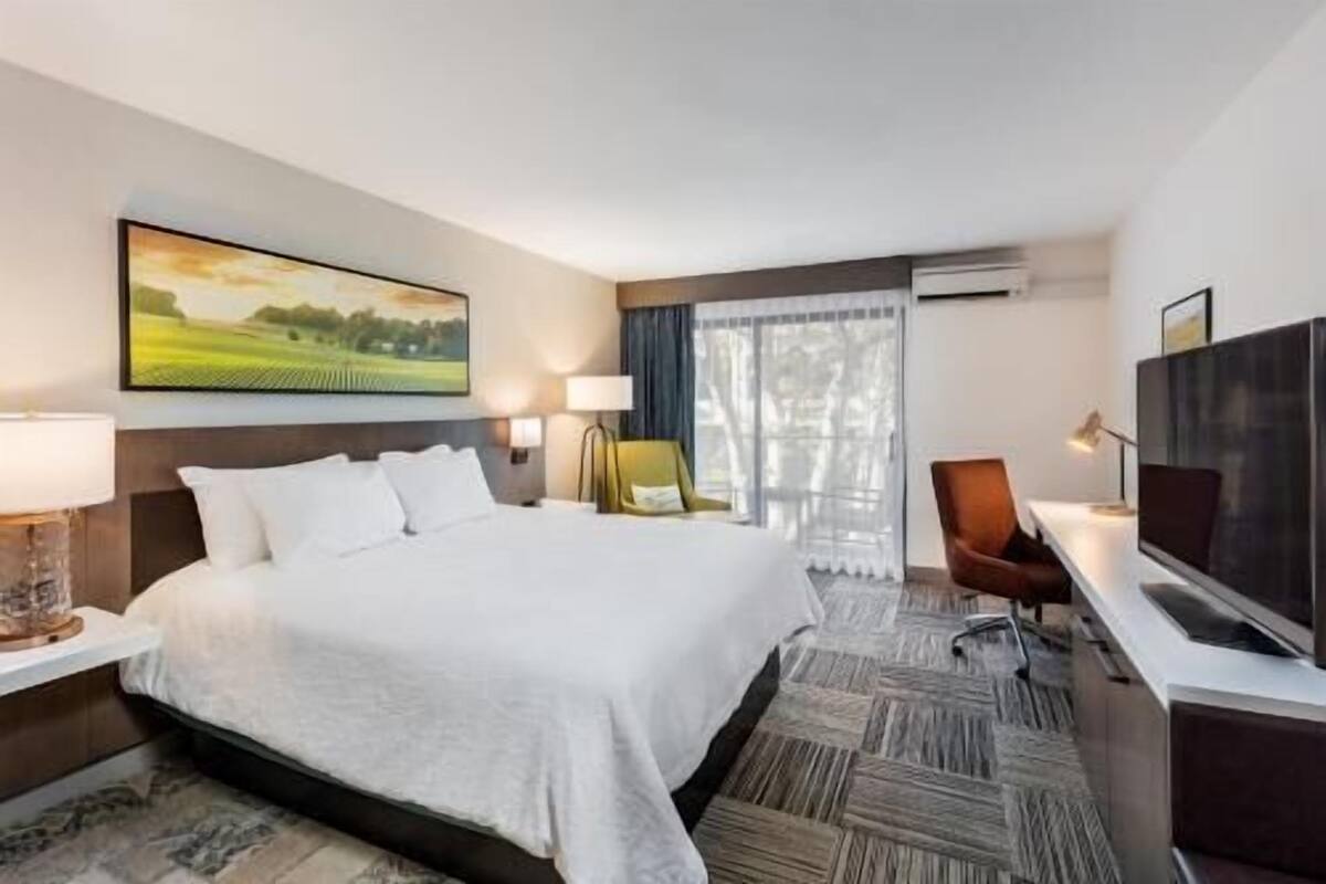 A hotel room with a large bed, a desk with a TV, a chair, and a window with curtains.