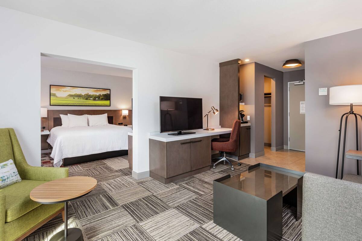 A modern hotel room with a bed, a desk with a TV, a green armchair, a glass coffee table, and a view of a field.