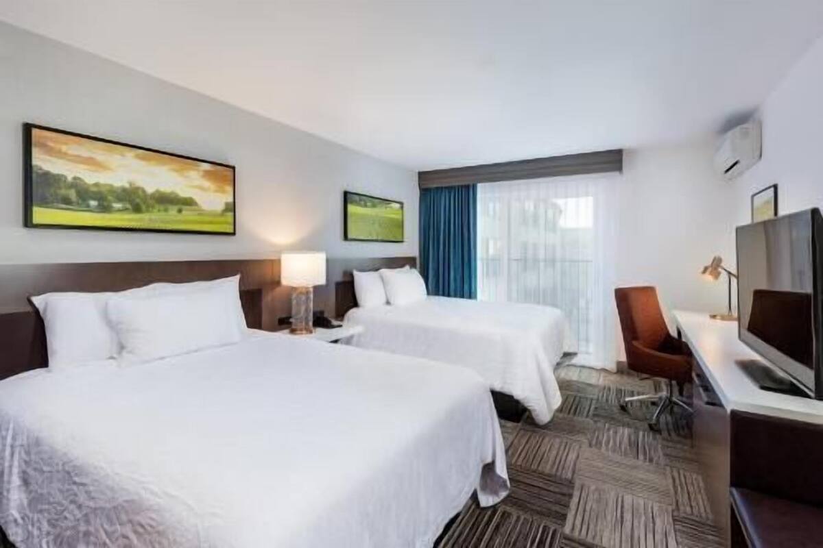 A hotel room with two beds, a desk with a chair, a television, and two paintings on the wall.