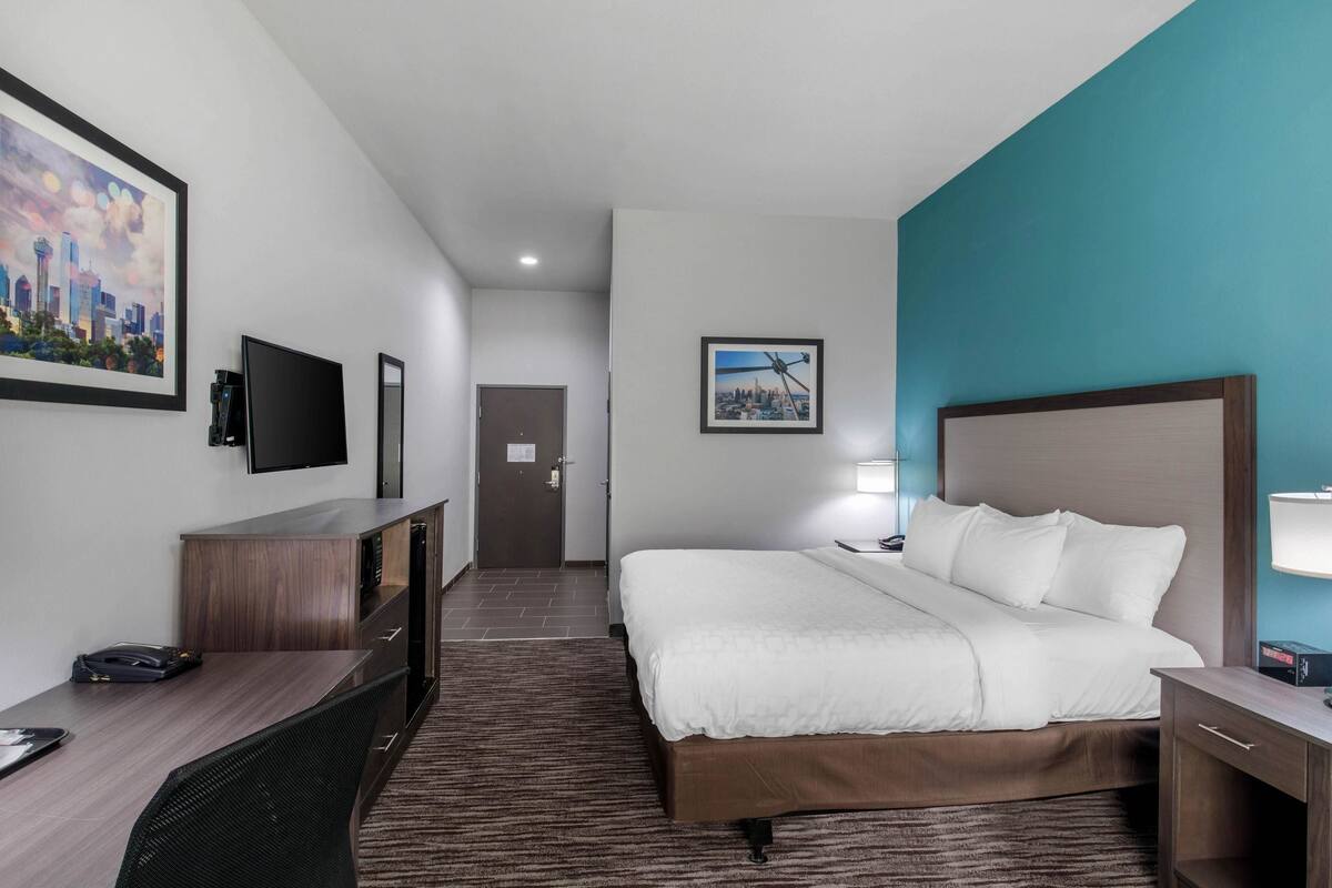A hotel room with a large bed, a desk with a computer, a TV mounted on the wall, and a door leading to another room.