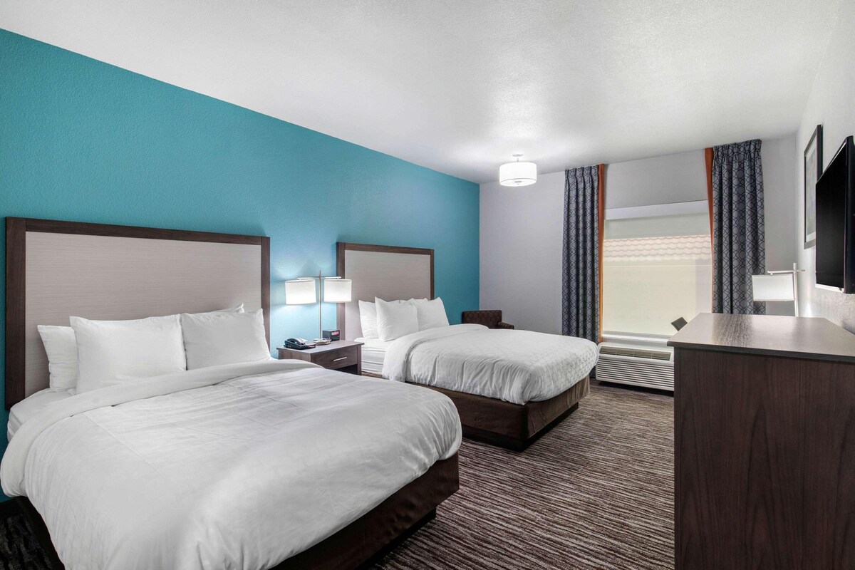 Quarto de hotel com duas camas, uma escrivaninha, uma cadeira, televisão e janela com cortinas.
