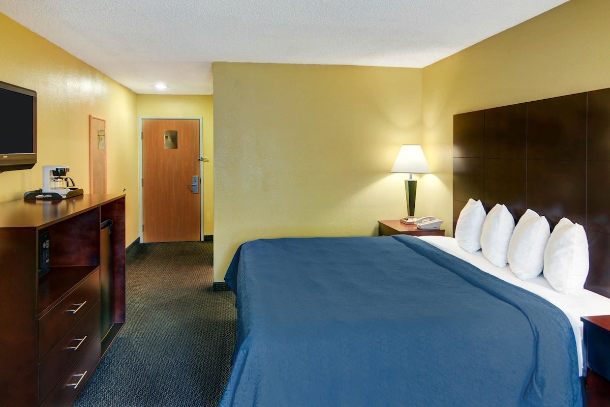 A hotel room with a bed, a desk with a lamp, a television, and a door.