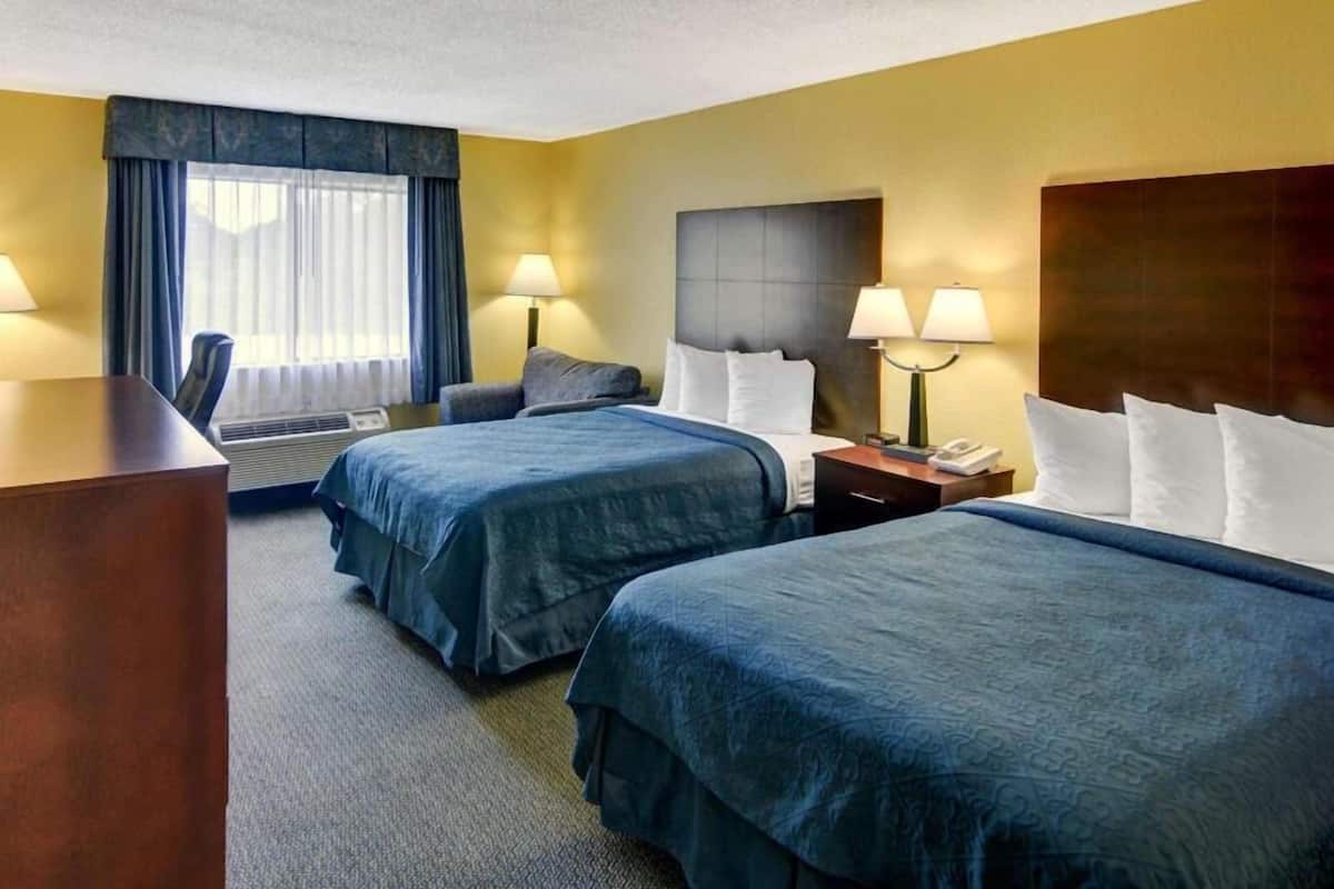 A hotel room with two beds, a desk, a chair, a lamp, and a window with curtains.