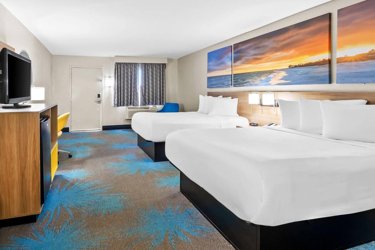A hotel room with two beds, a desk with a computer, a TV, and a large window with a sunset view.