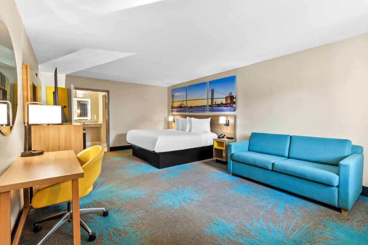 A hotel room with a bed, a desk, a yellow chair, a blue sofa, and a painting of a bridge.