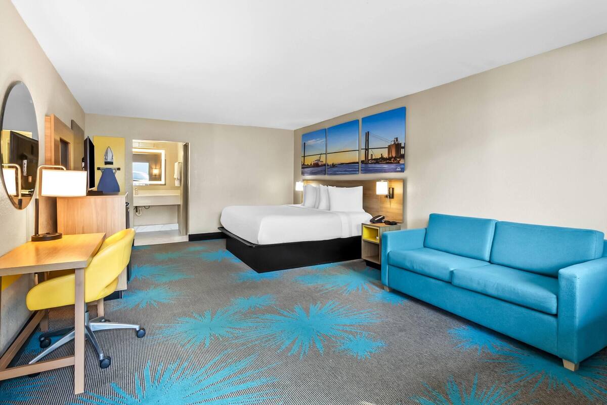 A hotel room with a bed, a blue sofa, a desk with a chair, a mirror, and a painting of a bridge.
