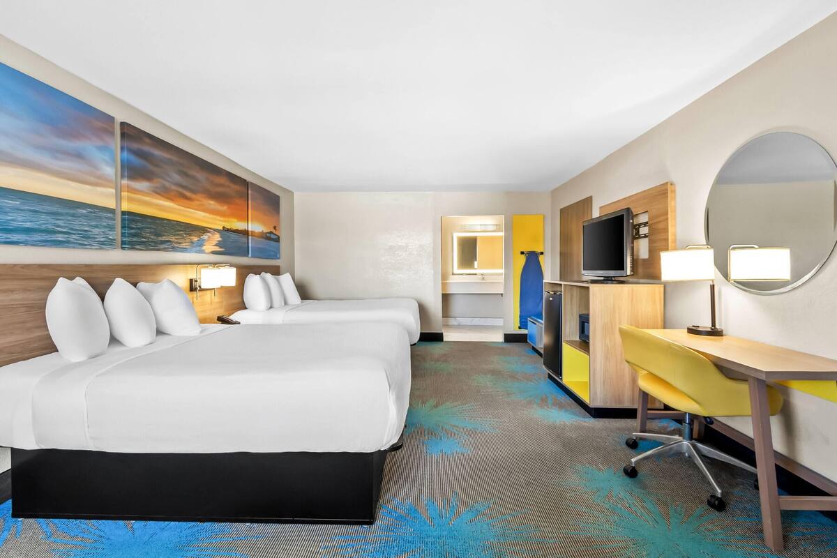 A hotel room with two beds, a desk, a TV, and a large painting of a sunset.