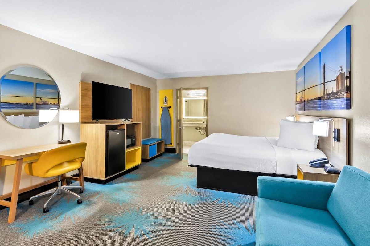 A hotel room with a bed, desk, chair, TV, and a large mirror.