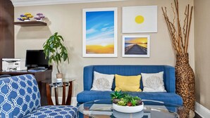 A living room with a blue sofa, a glass coffee table with plants, a TV, and artwork on the wall.