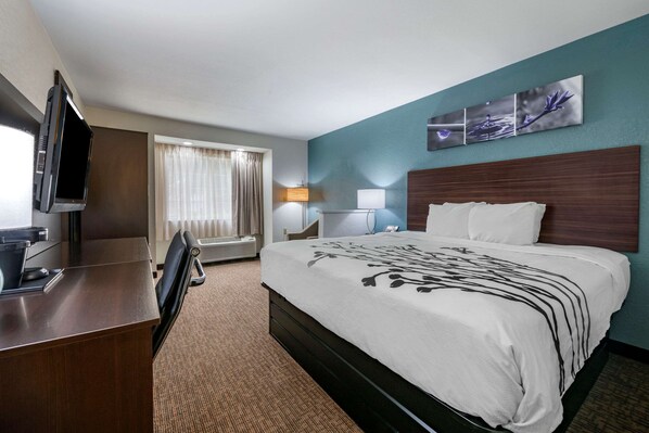 Una habitación de hotel con cama, escritorio, televisión, lámpara y ventana con cortinas.