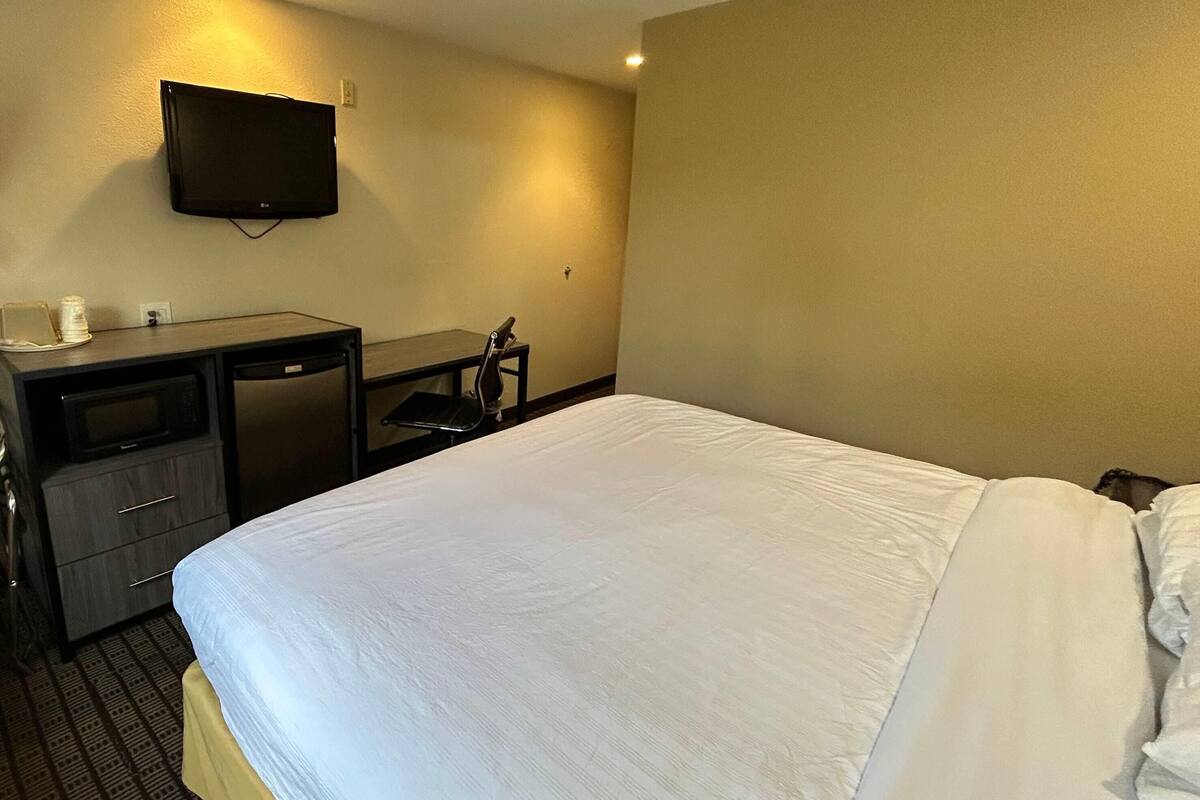 Ett hotellrum med en säng, ett skrivbord, en stol, en tv som är monterad på väggen och ett litet bord med en kopp och ett fat.