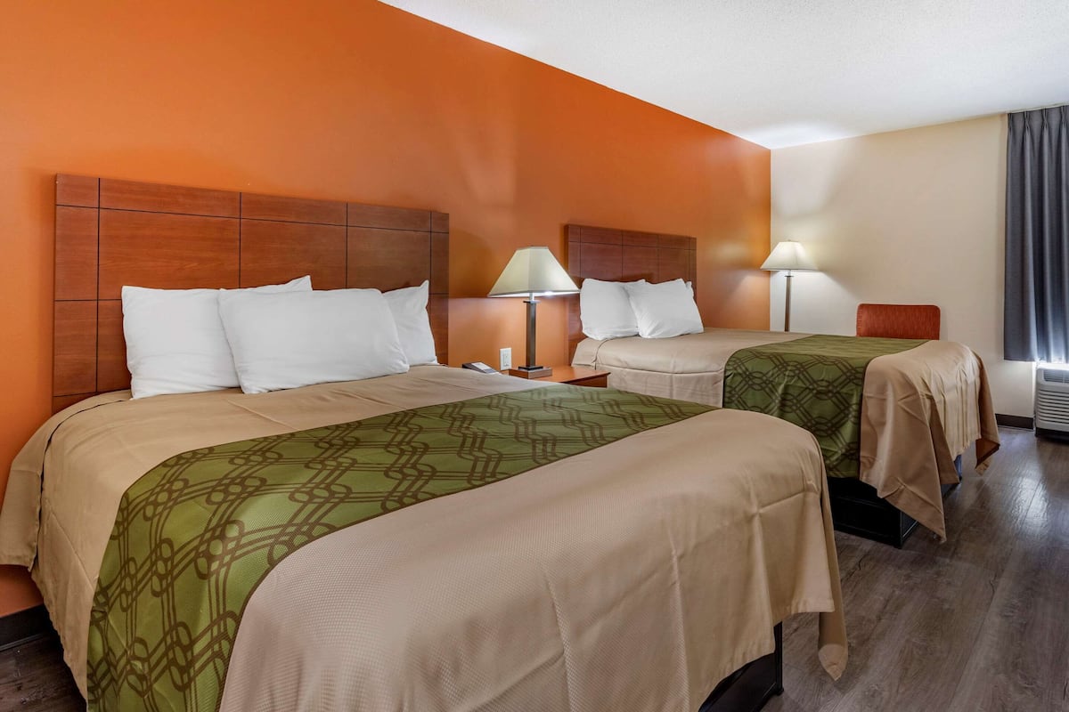 Une chambre d’hôtel avec deux lits, une tête de lit en bois, une table de chevet avec une lampe et une fenêtre avec des rideaux.