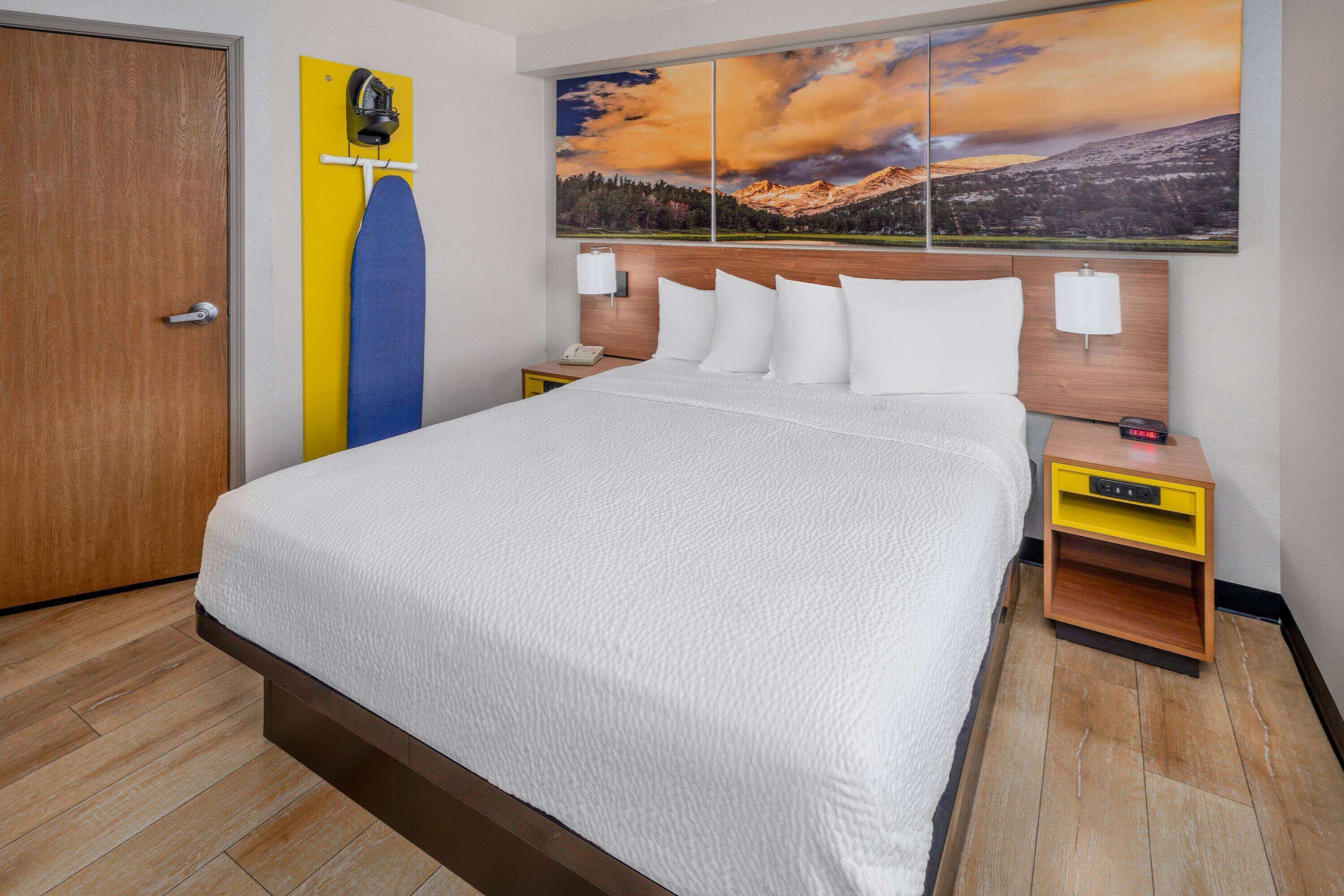 Habitación de hotel con una cama grande, cabecera de madera, un cuadro de un paisaje montañoso, una mesita de noche amarilla con lámpara y una puerta de madera.