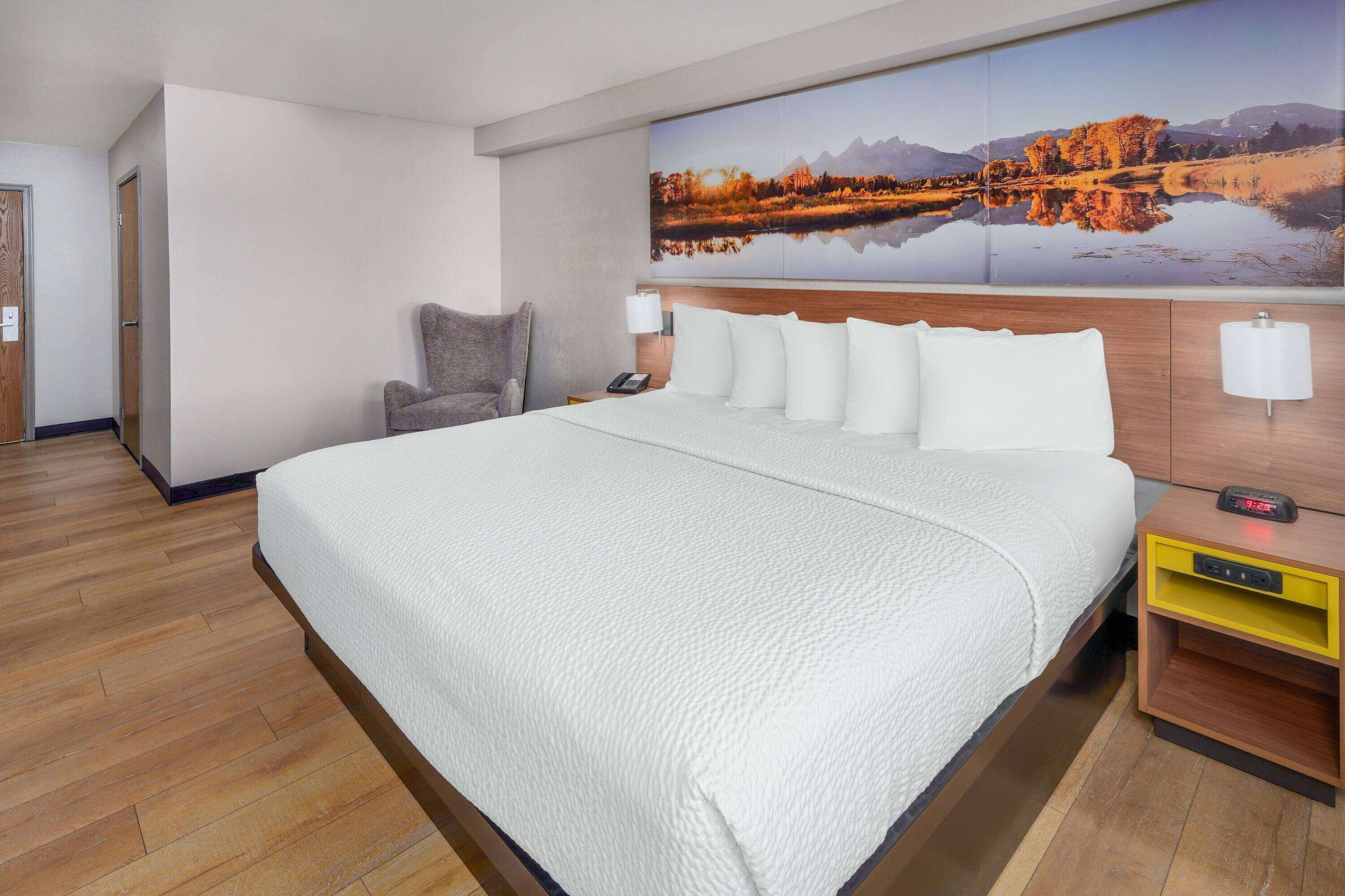 A hotel room with a large bed, a chair, a nightstand with a lamp, and a wall-mounted painting of a landscape.