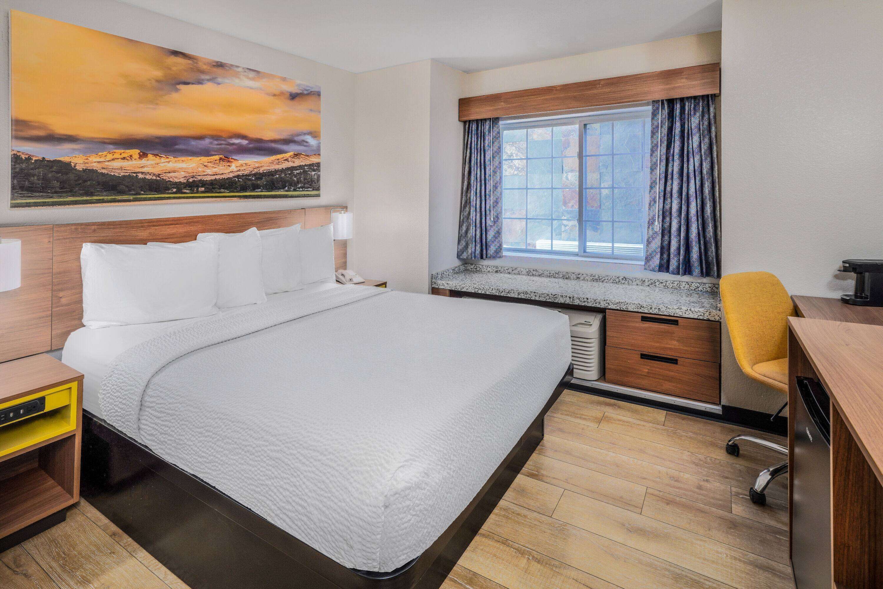 A hotel room with a large bed, a desk, a chair, a window with curtains, and a painting on the wall.