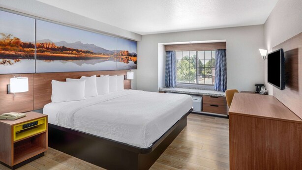 A hotel room with a large bed, a desk, a TV, a window with curtains, and artwork on the wall. - Days Inn & Suites by Wyndham Greeley (Greeley)
