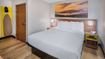 A hotel room with a bed, a wooden floor, a painting of a mountain landscape, and a yellow ironing board.