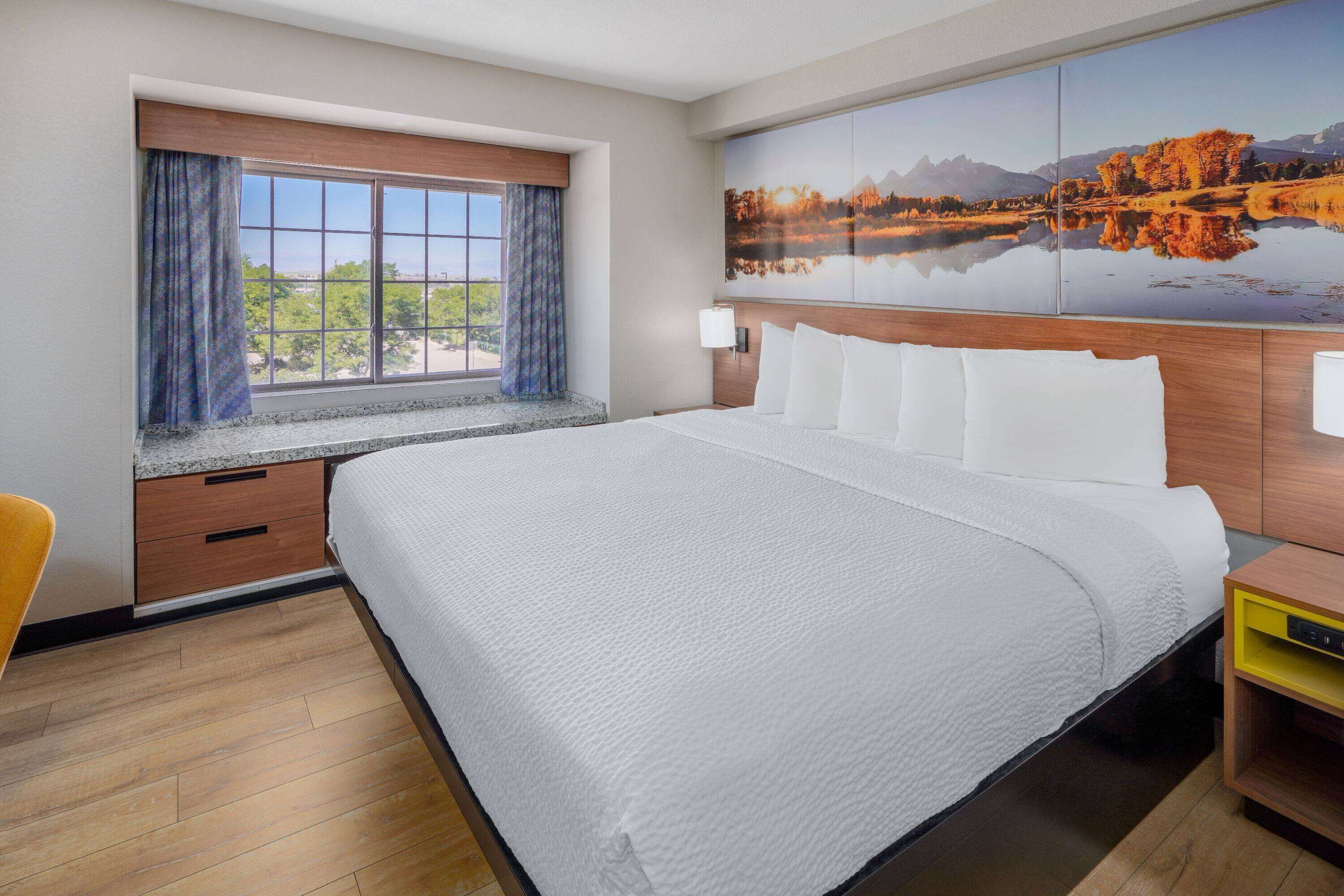 A hotel room with a large bed, a window with curtains, a painting of a landscape, and a wooden nightstand.