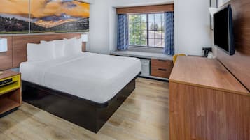 Quarto de hotel com cama, escrivaninha, televisão, janela com cortinas e um quadro na parede.