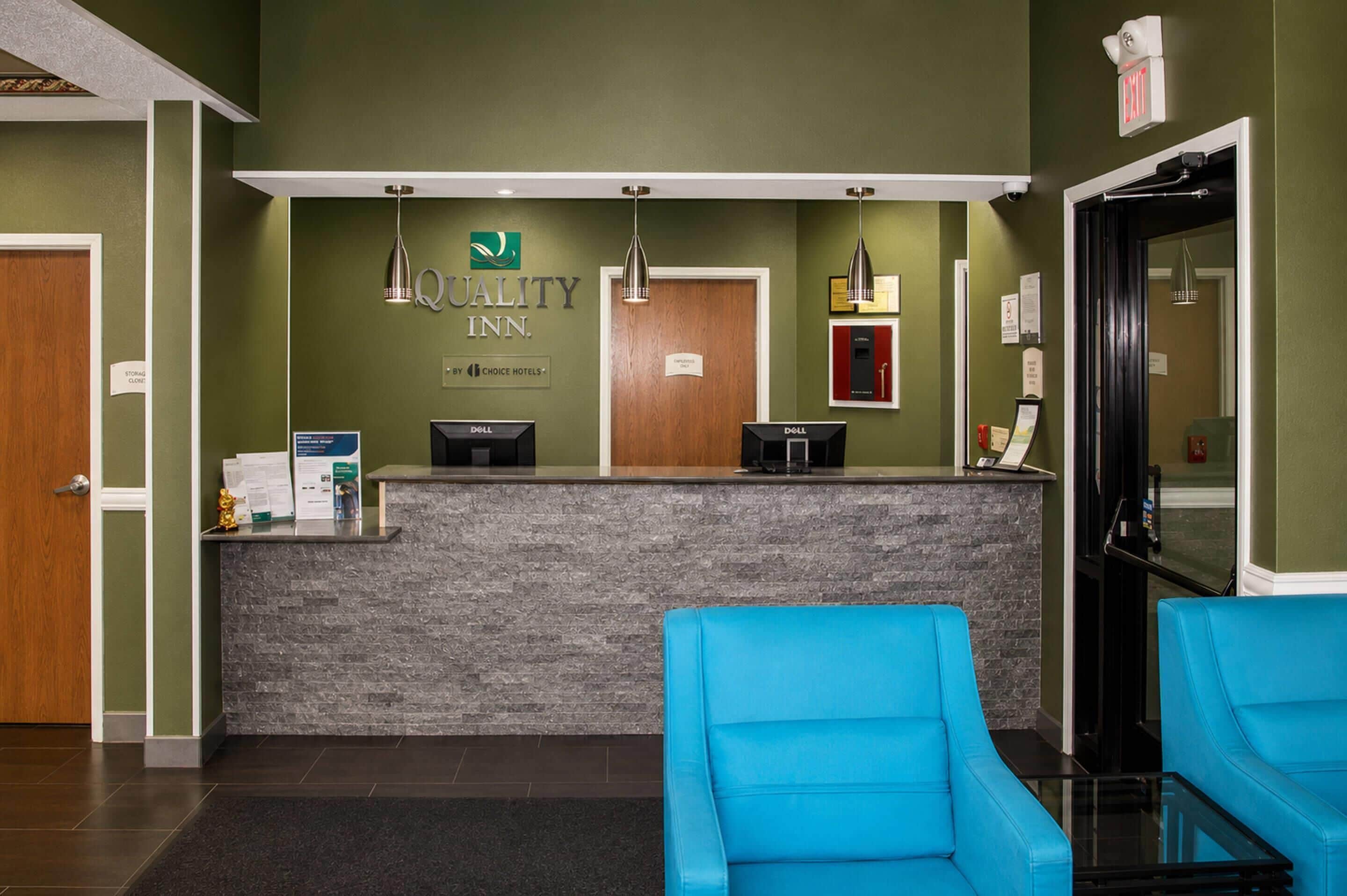 A hotel reception area with a reception desk, two blue chairs, and a wall-mounted sign for Quality Inn.