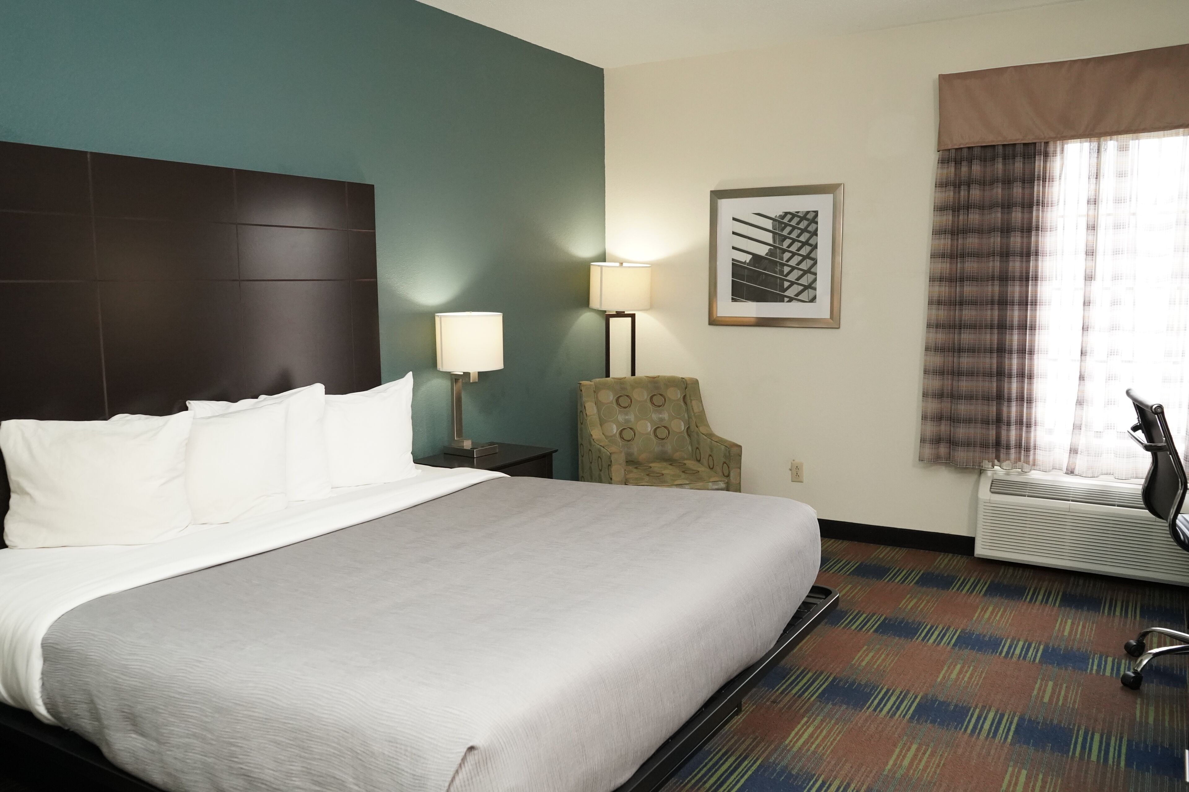 A hotel room with a large bed, a chair, a desk, a lamp, a framed picture, and a window with curtains.
