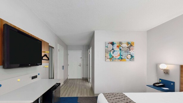 A hotel room with a flat-screen TV, a desk with a computer, a bed with patterned bedding, a wall art piece, and a bedside table with a lamp. - Microtel Inn by Wyndham Bowling Green (Bowling Green)
