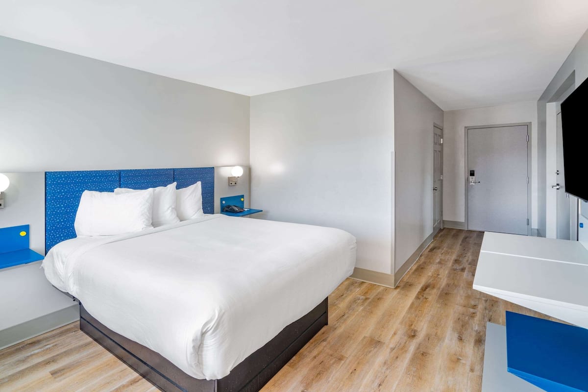 A modern hotel room with a large bed, a bedside table, a nightstand, a TV, and a bathroom with a white sink and mirror.
