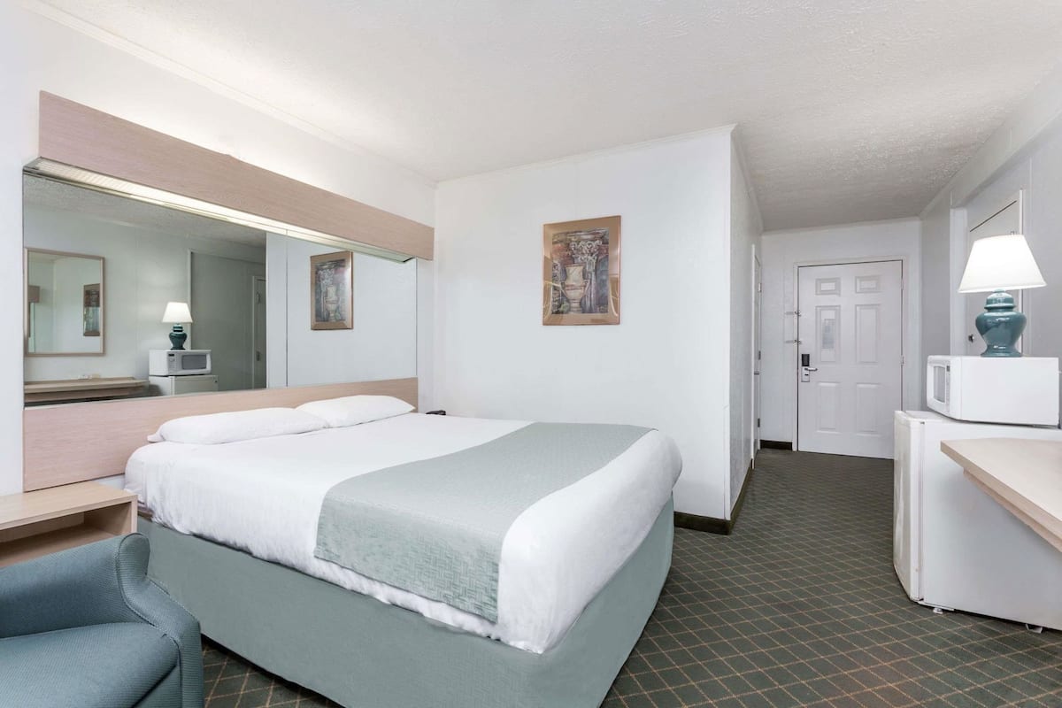 A hotel room with a bed, a desk, a chair, a mirror, a door, and a hallway.