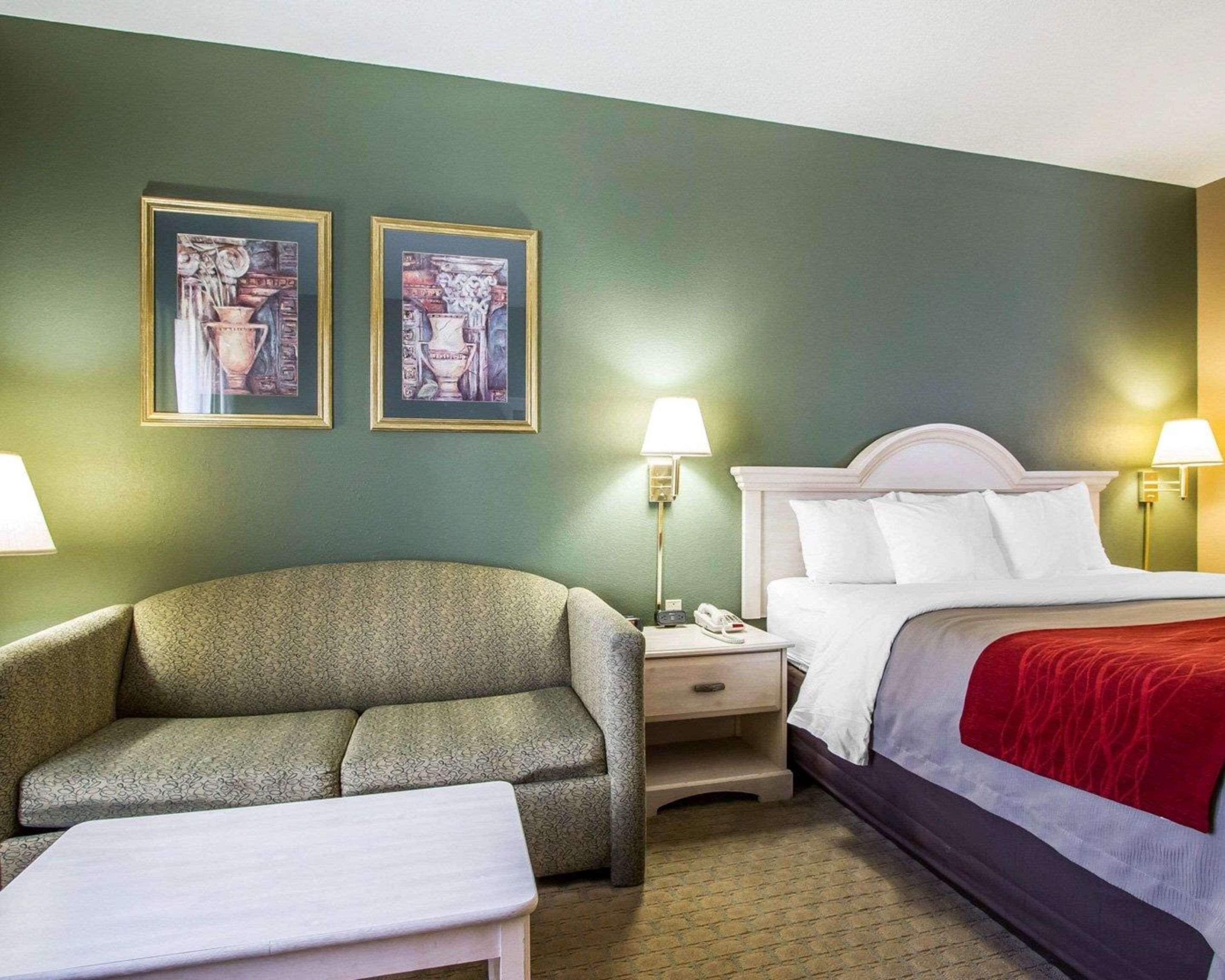 Une chambre d’hôtel avec un mur vert, deux œuvres d’art encadrées, un canapé, un lit avec une literie blanche et un plaid rouge, ainsi que des lampes de chevet.