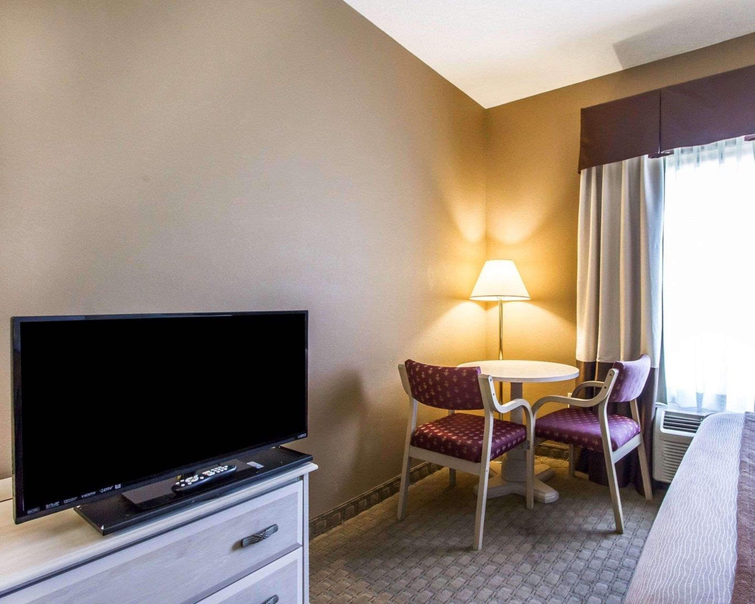 A hotel room with a flat-screen TV on a dresser, a small round table with two chairs, a lamp, and a window with curtains.