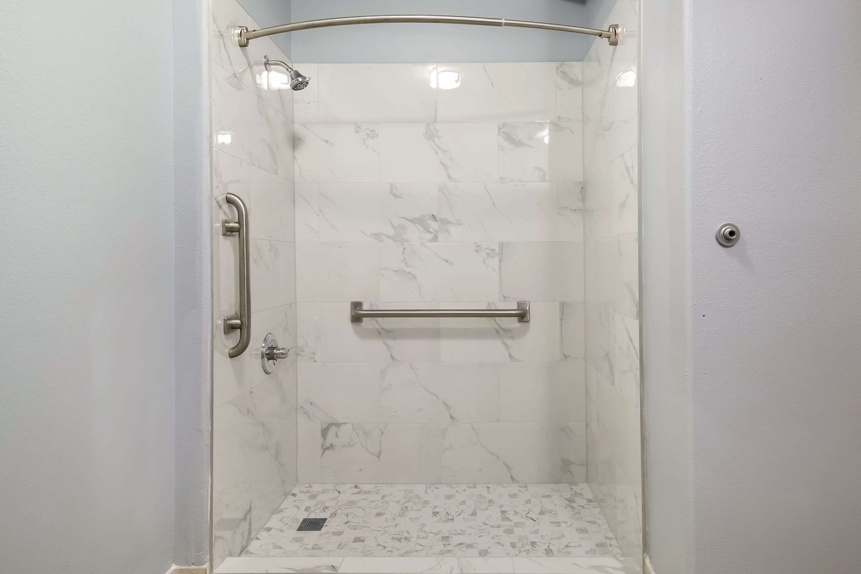 Ducha con puerta de vidrio, azulejos de mármol, barra de apoyo y toallero.