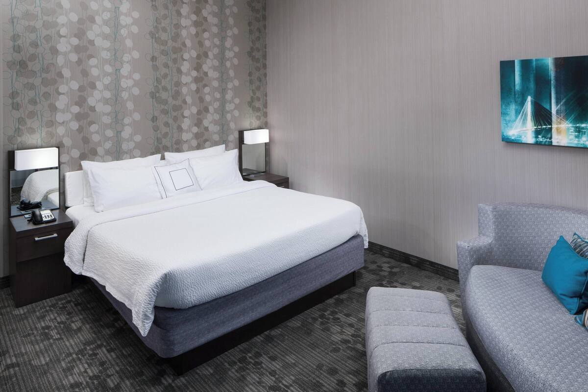 A hotel room with a large bed, two bedside tables with lamps, a grey sofa, and a wall-mounted painting.