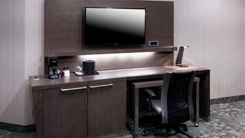 A modern hotel room with a flat-screen TV mounted on a wooden wall, a desk with a chair, a coffee maker, and a lamp.