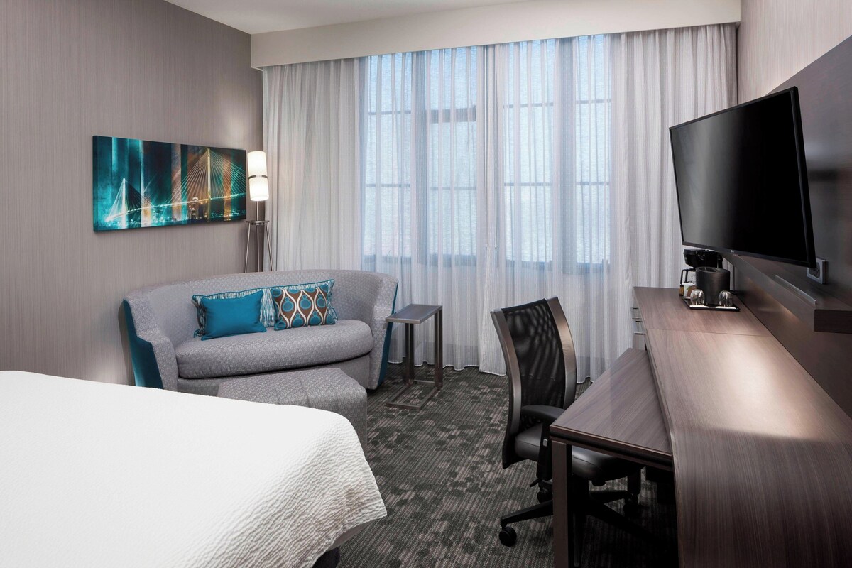 A hotel room with a bed, a gray sofa with a patterned pillow, a small side table, a desk with a flat-screen TV, and a large window with sheer curtains.