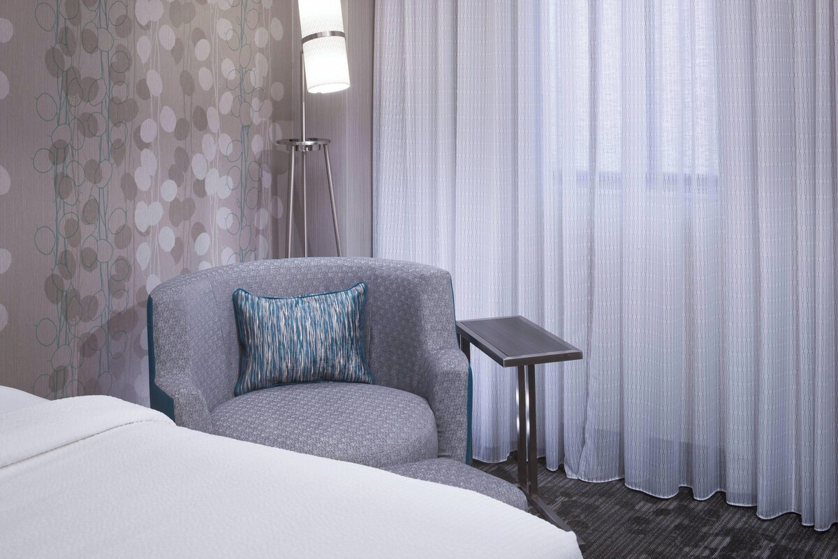 A hotel room with a grey armchair, a small table, a white bed, and a view through sheer curtains.