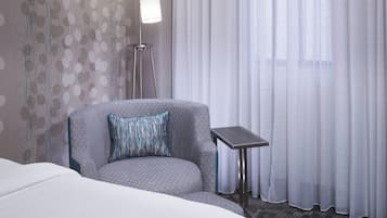 Habitación de hotel con un sillón gris, una mesita pequeña, una cama blanca y vistas a través de cortinas translúcidas.