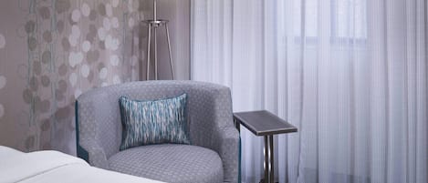 Habitación de hotel con un sillón gris, una mesita pequeña, una cama blanca y vistas a través de cortinas translúcidas.