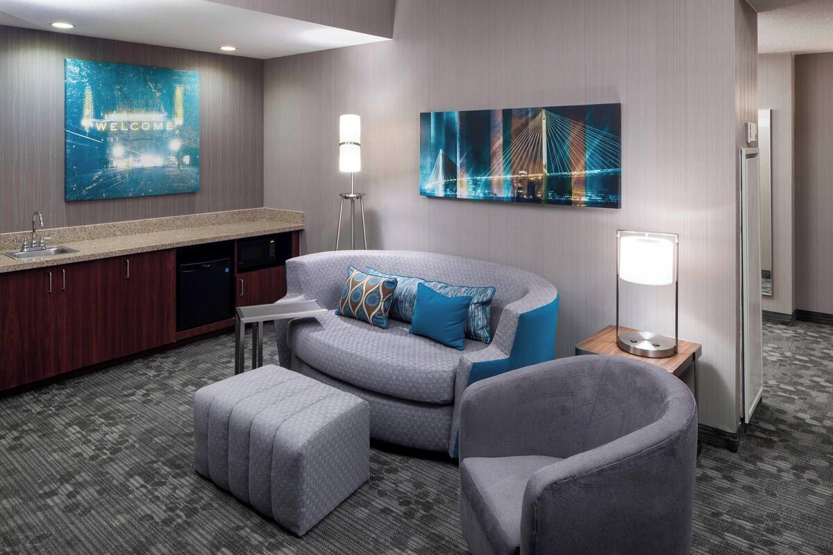 A modern hotel room with a kitchenette, a sofa, a chair, and a desk.