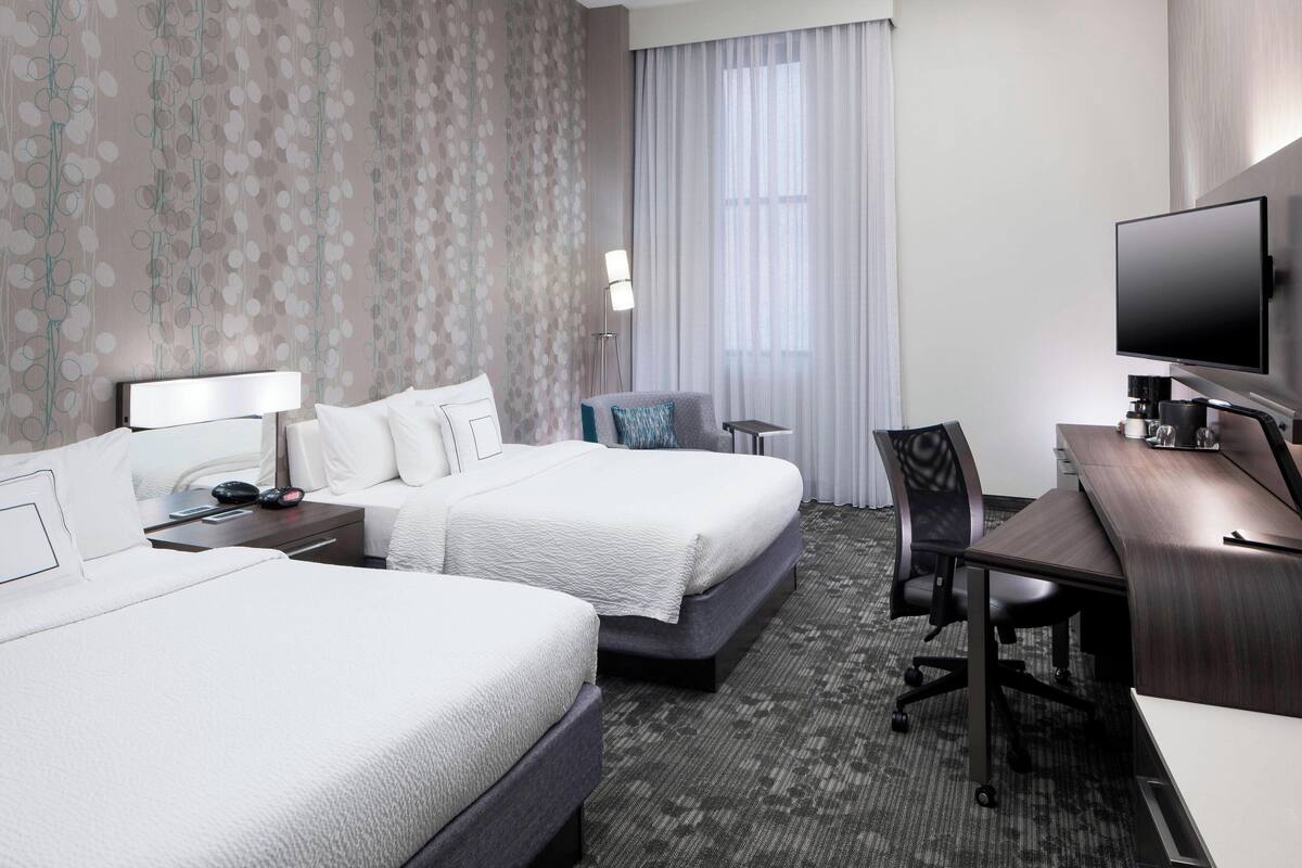 A hotel room with two beds, a desk, a chair, a television, and a lamp.