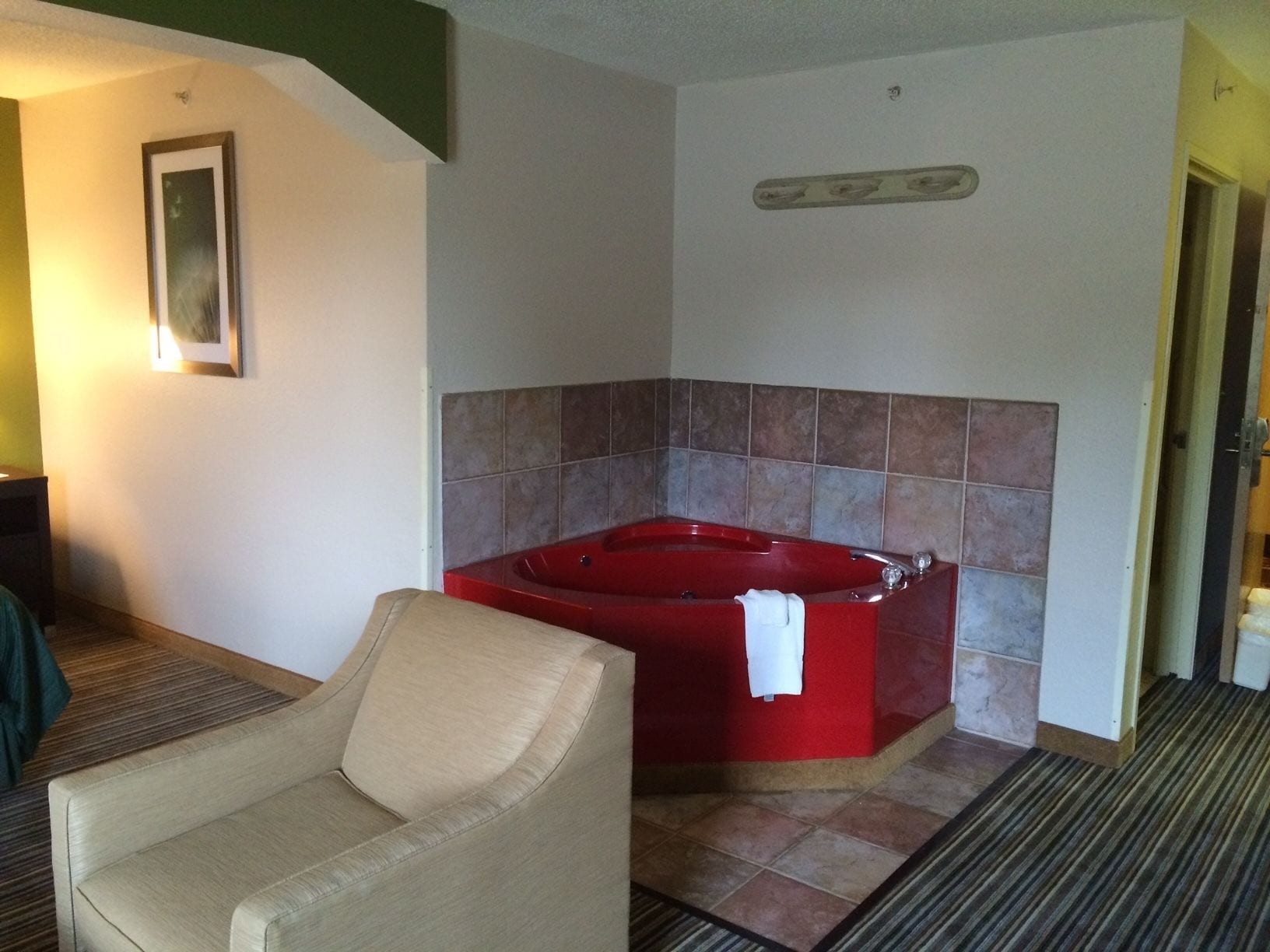 Une chambre d’hôtel avec un jacuzzi rouge, un fauteuil beige et un tableau encadré au mur.