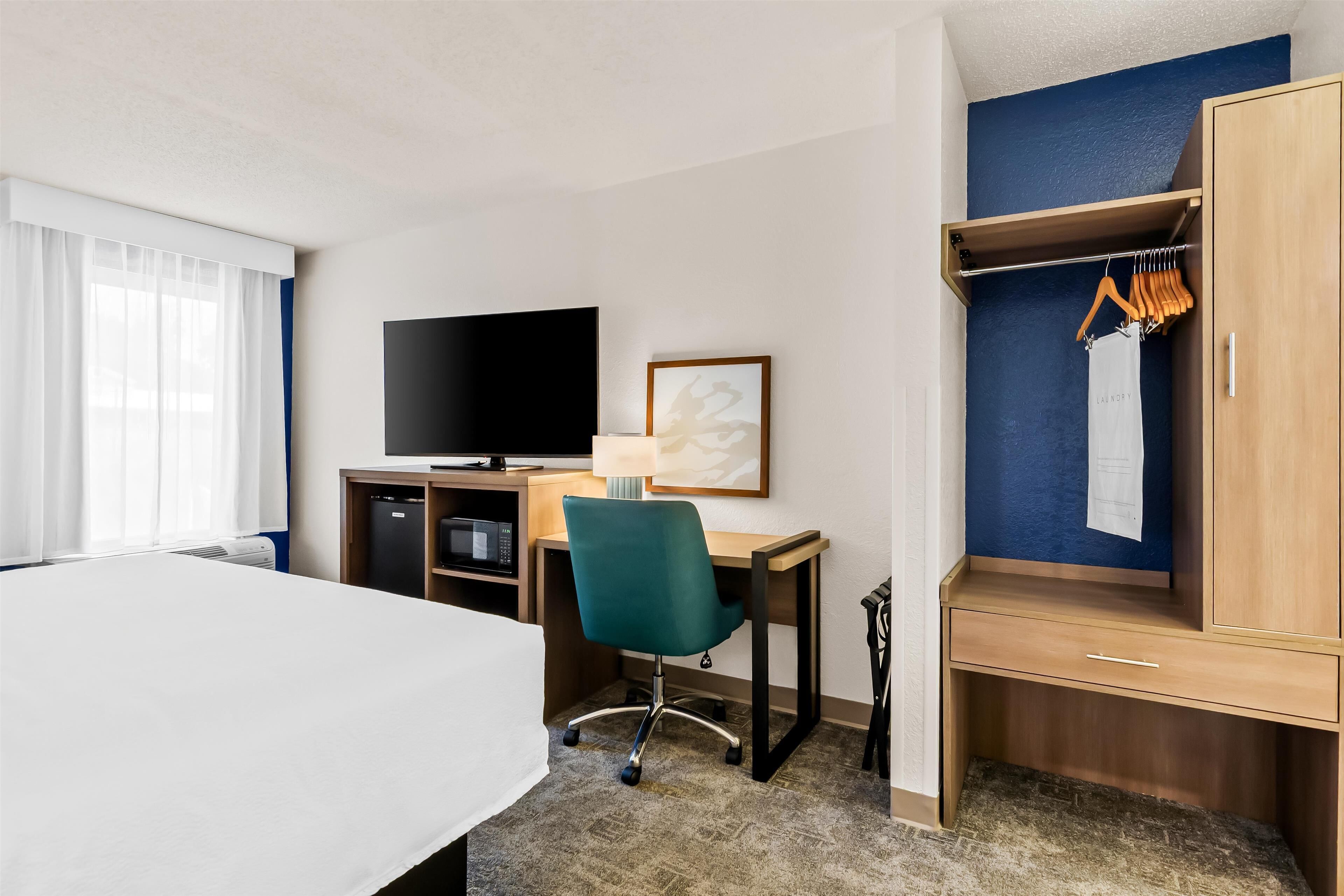 A hotel room with a bed, desk, chair, TV, wardrobe, and a painting on the wall.