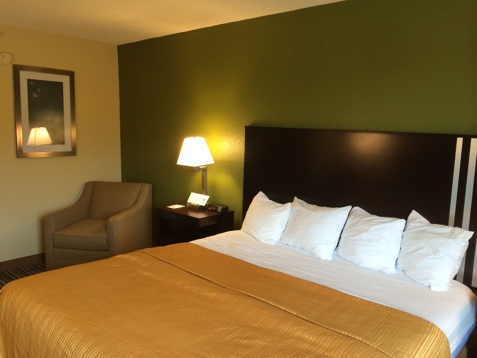 Habitación de hotel con una cama grande, una mesita de noche con lámpara, una silla e imagen enmarcada en la pared.